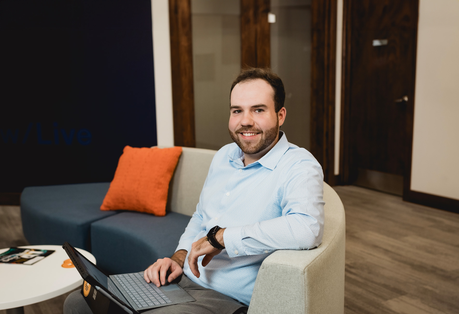Ben Petell, Associate Financial Advisor: A man with short brown hair and a beard wearing a blue shirt. Working on his computer sitting on a couch. Strategic Advisor Team bio page headshot.