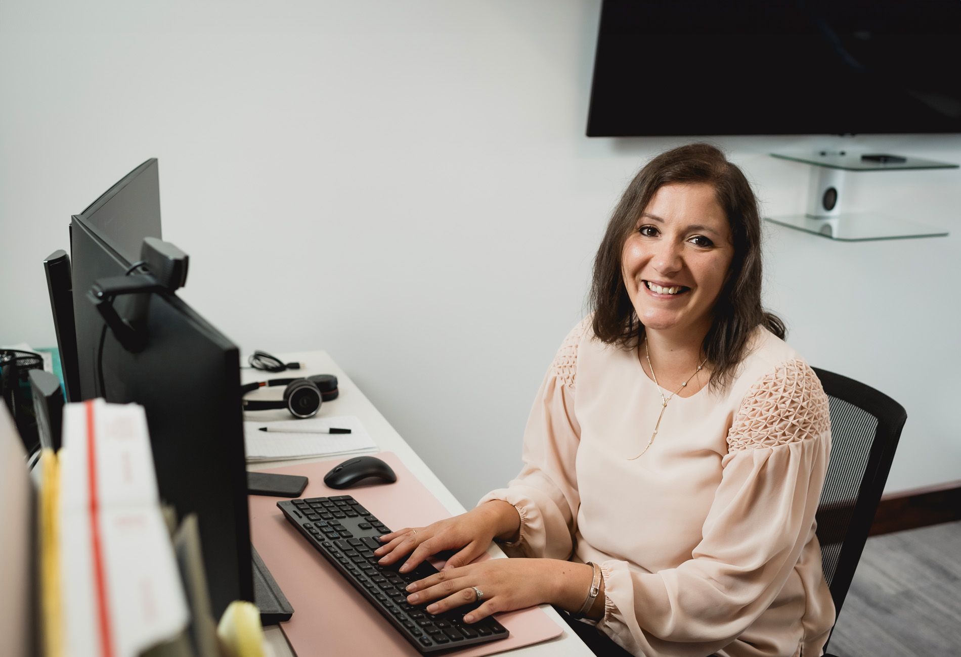 Holly Cafalone, Operations Manager: A woman with shoulder length brown hair in a pink blouse working at her computer. company headshot photo.