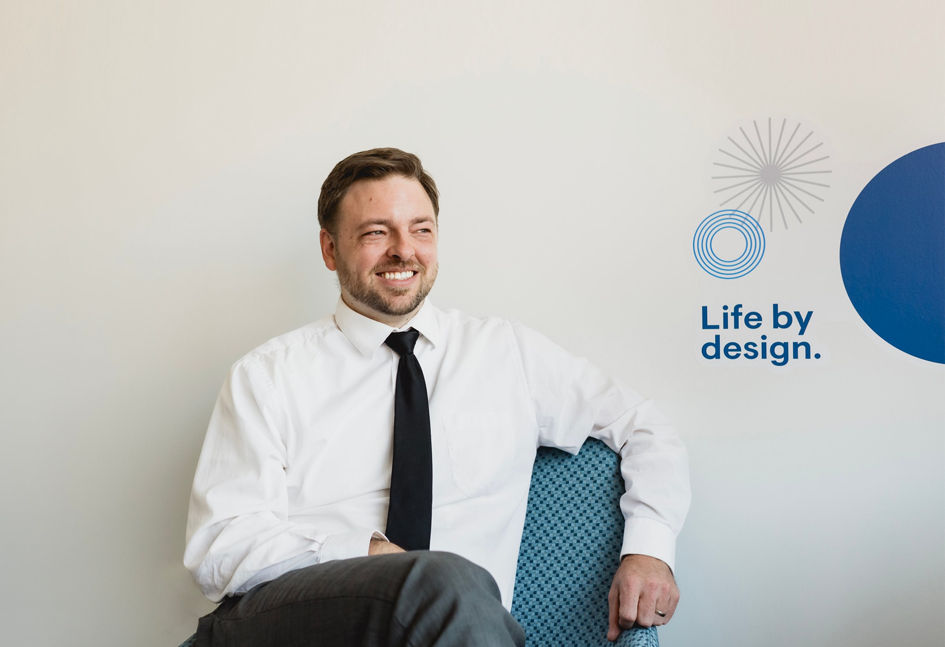 Kyle Dempster, CFP, Lead Financial Advisor: A man with short brown hair and a short beard wearing a white button down and tie against a white wall with a company tagline – life by design.