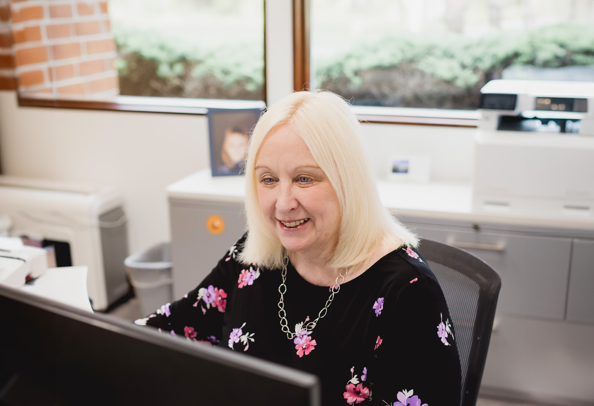 Nancy Meininger, Head of Business Management: A woman with long blonde hair and a black and floral blouse working at her computer.
