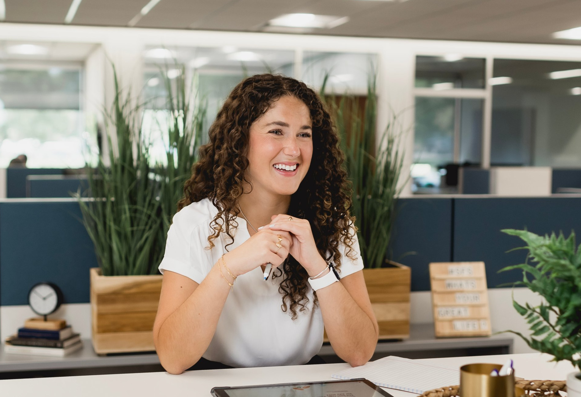 Sophia Tsoupelis, Associate Financial Advisor: A young woman with brown curly hair wearing a white blouse and sitting at a work table.